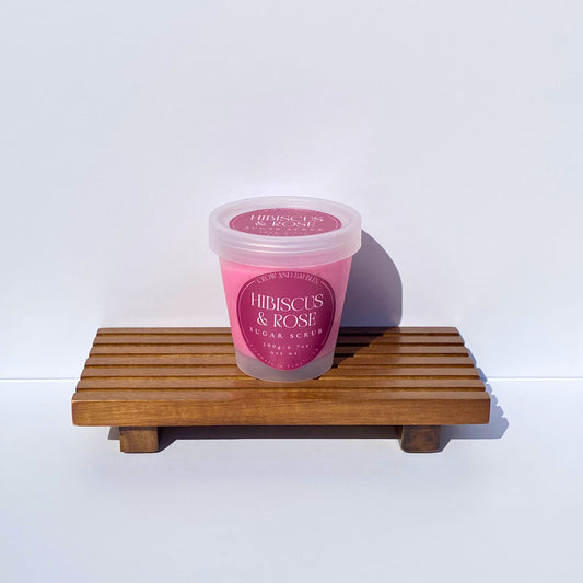 Pink jar of 'Hibiscus & Rose' sugar scrub on a wooden stand with a white background