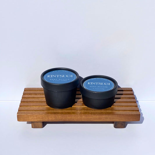 Two black containers with blue Kintsugi labels on a wooden stand against a white background