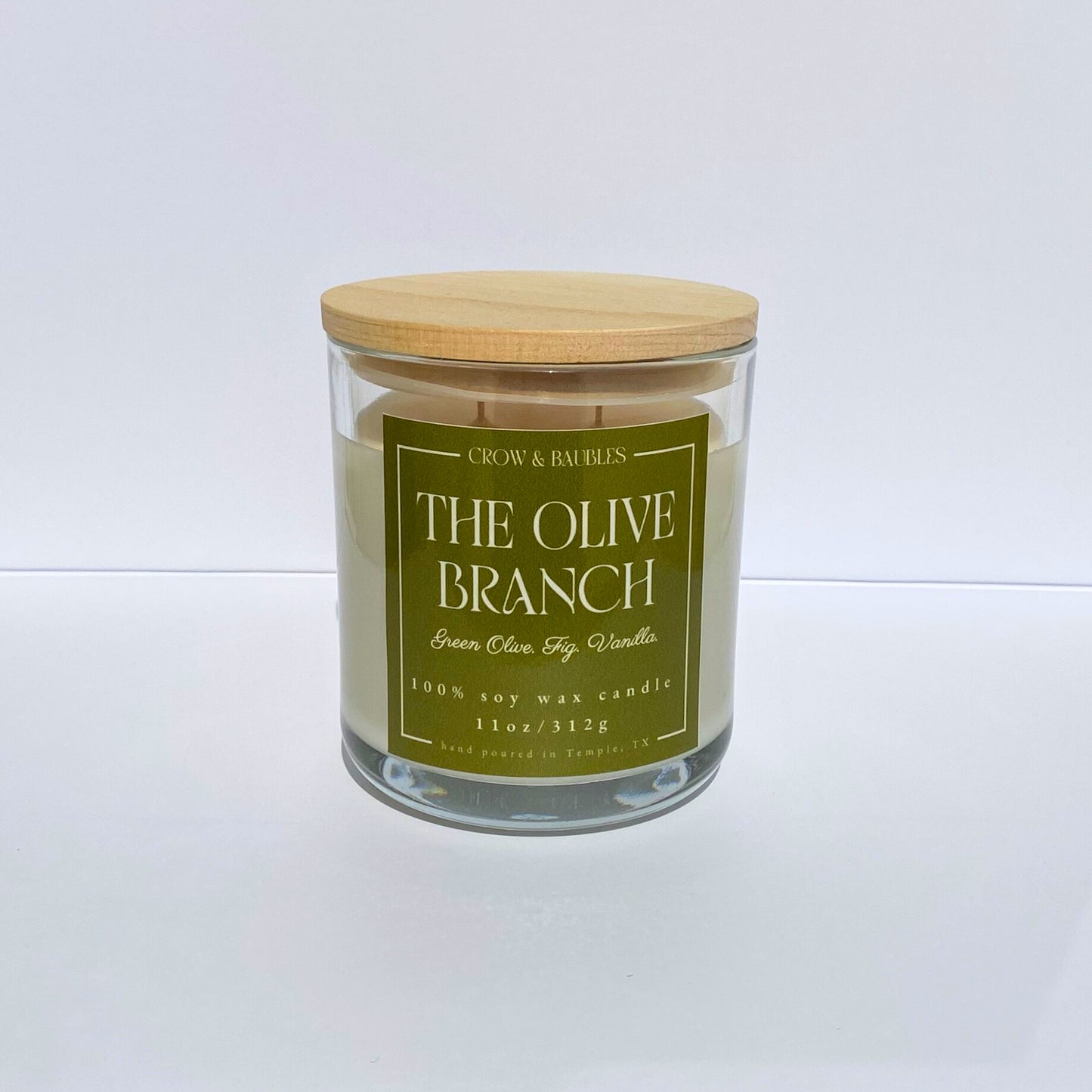 Candle in a glass jar with a wooden lid and green label "The Olive Branch" on a white background