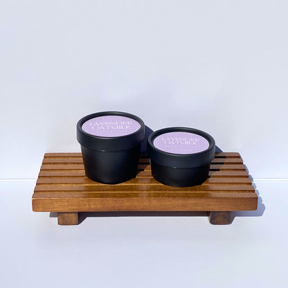 Two black containers with lavender labels on a wooden stand against a white background