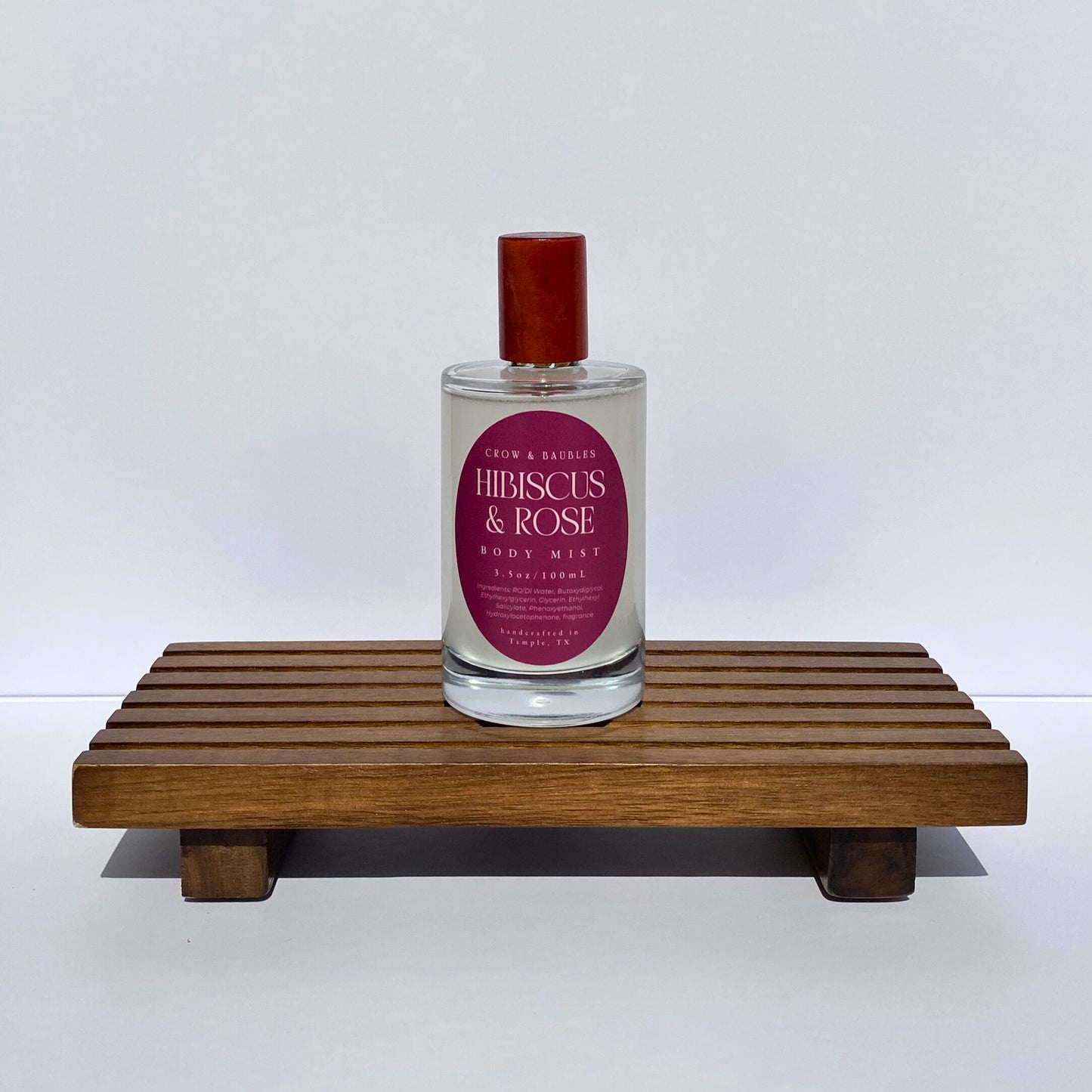 Bottle of 'Hibiscus & Rose' body mist on a wooden stand with a white background