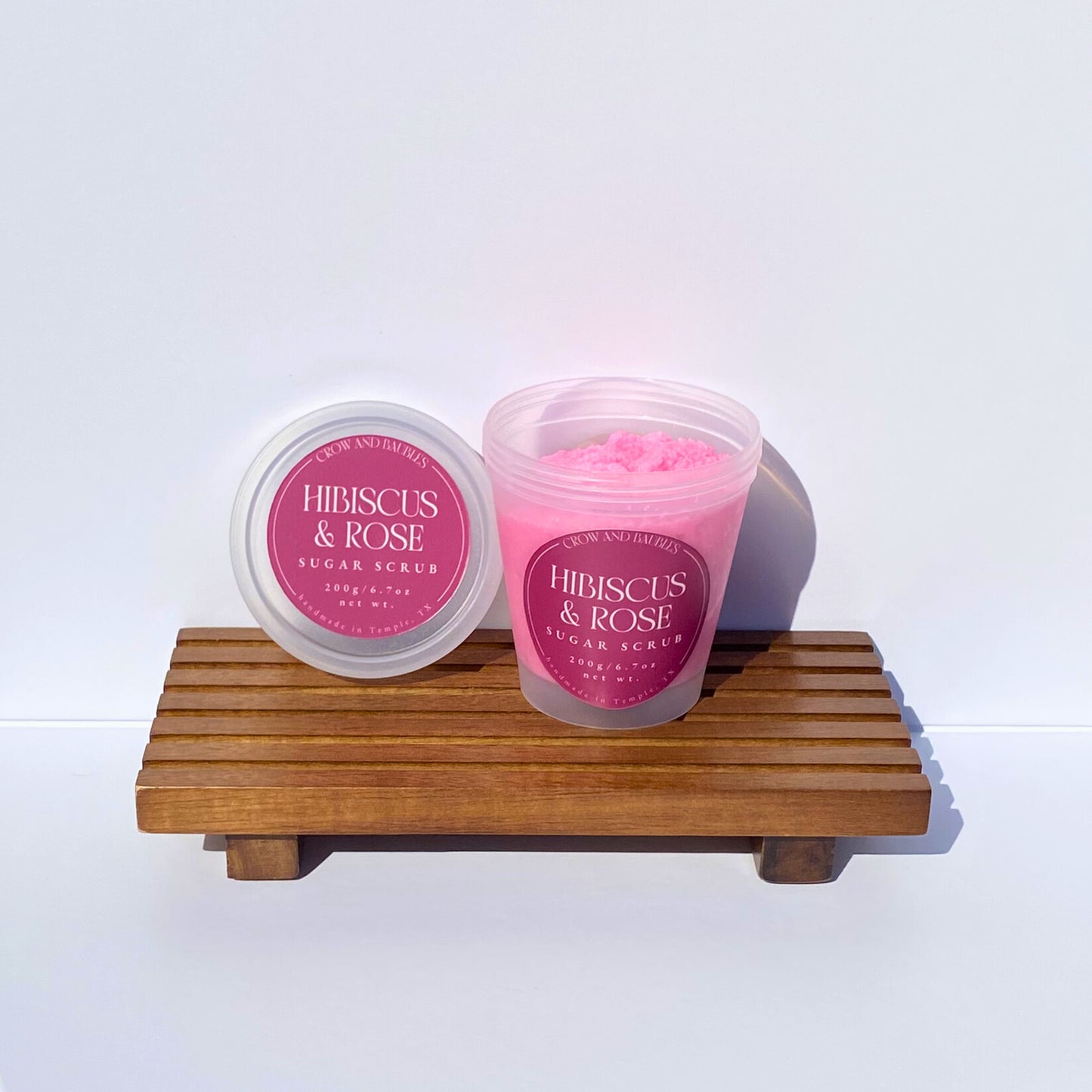 an open container of Hibiscus & Rose sugar scrub on a wooden stand with a white background