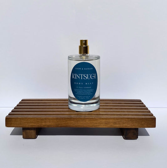 Clear spray bottle labeled 'Kintsugi' on a wooden stand with a white background