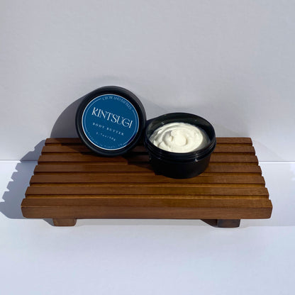 Two containers of Kintsugi body butter on a wooden stand with a white background