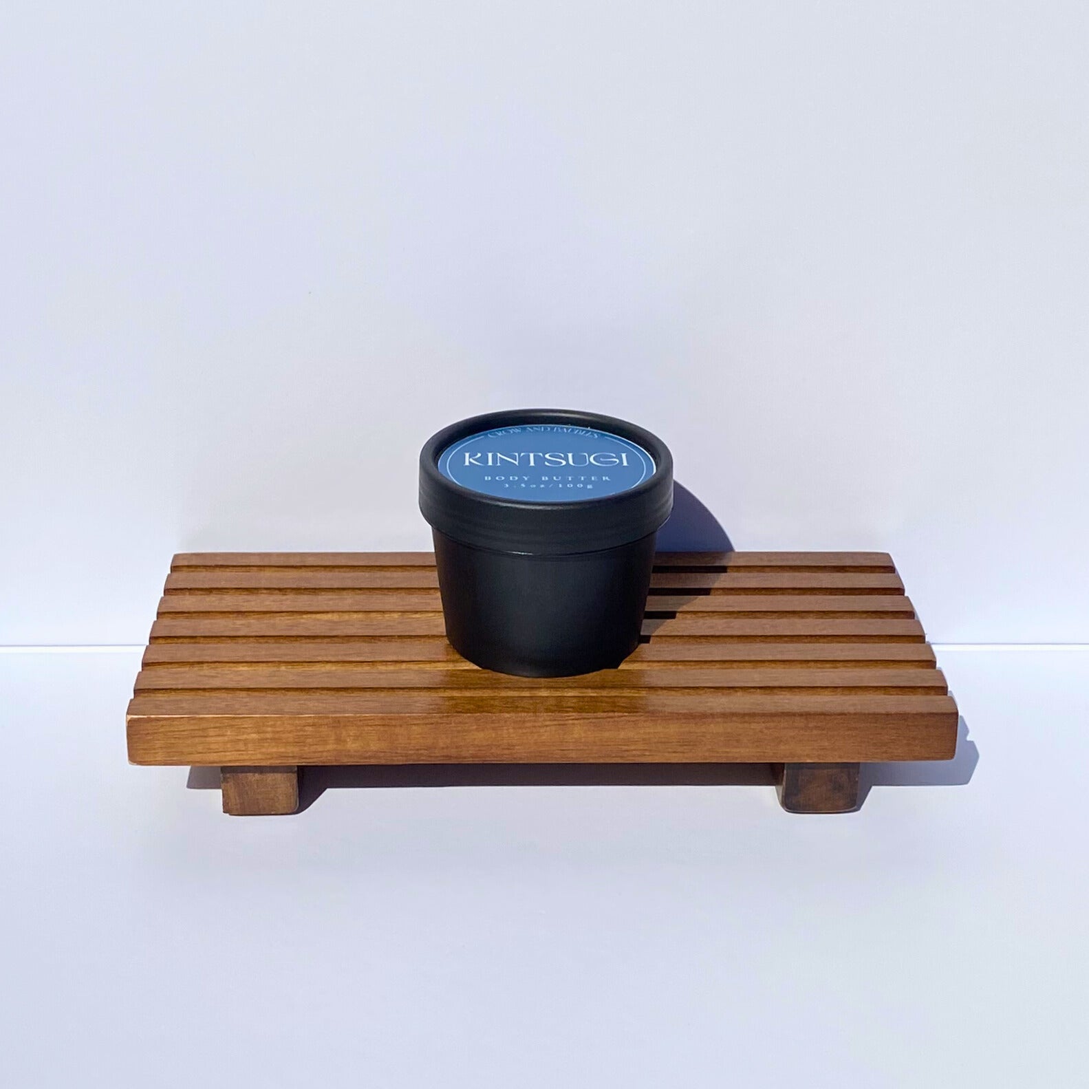 Black container with blue Kintsugi label on a wooden tray against a white background