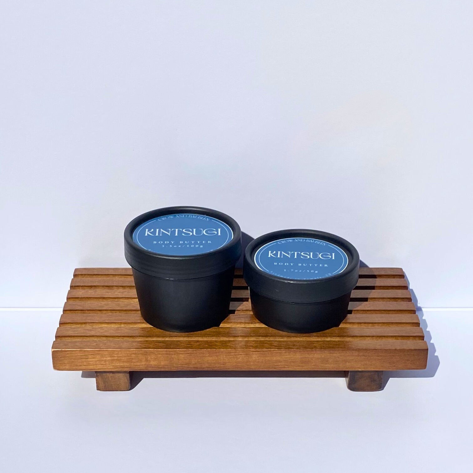 Two black containers with blue Kintsugi labels on a wooden stand against a white background