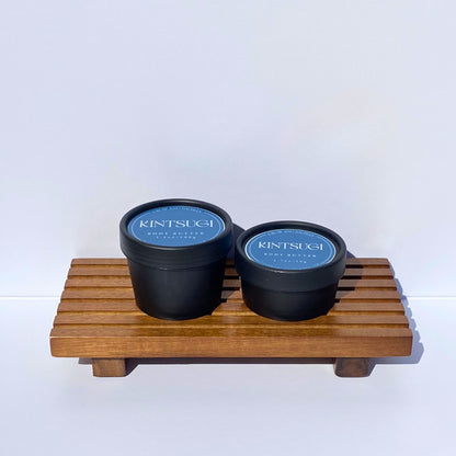 Two black containers with blue Kintsugi labels on a wooden stand against a white background